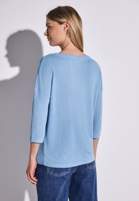 Helles blaues, weites Sweatshirt mit Dreiviertelärmeln und weitem Ausschnitt, aus einem strukturierten Stoff gefertigt. Kombiniert mit Jeans.