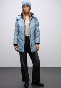 Lichtblauwe pufferjack met ruitpatronen, zwarte voering, ritssluiting en zijzakken. Gecombineerd met zwarte wijde broek en een blauwe gebreide beanie.