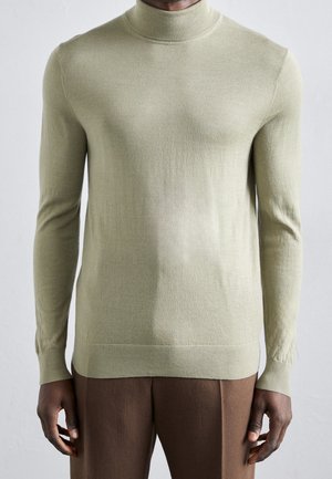 Homme portant un pull à col roulé beige clair et un pantalon marron, se tenant devant un fond uni gris clair.