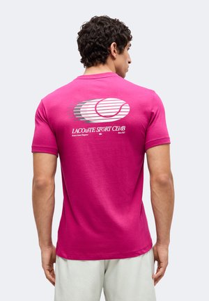Mężczyzna w jaskraworóżowej koszulce Lacoste Sport Club z białą grafiką piłki tenisowej i tekstem na plecach, w połączeniu z jasnymi szortami.