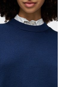 Sweater azul-marinho canelado com decote redondo, tecido texturizado, e uma camada de uma camisa de colarinho listrada branca e azul por baixo.