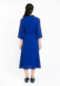 Plava haljina srednje dužine s opuštenim krojem, rukavima do tri četvrtine i dugmetom na manžeti. Prozirna tekstura s lepršavim dizajnom.