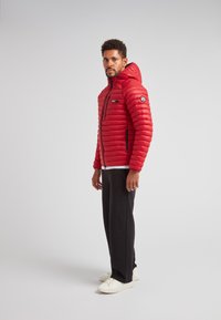 Giacca a vento trapuntata rossa con cappuccio, chiusura con zip e tasche laterali. Presenta un patch con logo sulla manica. Abbinata a pantaloni neri e sneakers bianche.