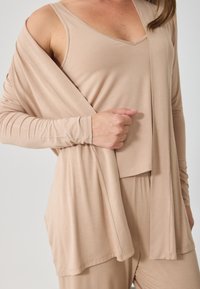 Η επένδυση από μπεζ ύφασμα περιλαμβάνει ένα μακρύ μανίκι καζάκα και ένα εφαρμοστό tank top. Απαλή υφή με χαλαρή σιλουέτα και διακριτές λεπτομέρειες ραφής.