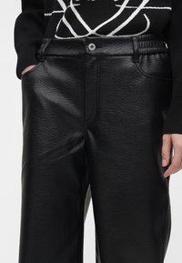 Gros plan sur un pantalon en cuir noir texturé avec bouton à l'avant et poches, porté avec un pull noir aux lignes abstraites blanches.