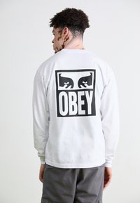 Long-sleeve white T-shirt featuring a large black "OBEY" logo and graphic on the back, made from soft cotton material.