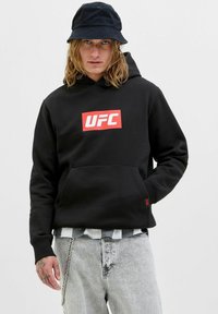 Persona con felpa nera UFC, jeans chiari, cappello a secchiello nero, mano sinistra tatuata e accessorio a catena sui jeans.