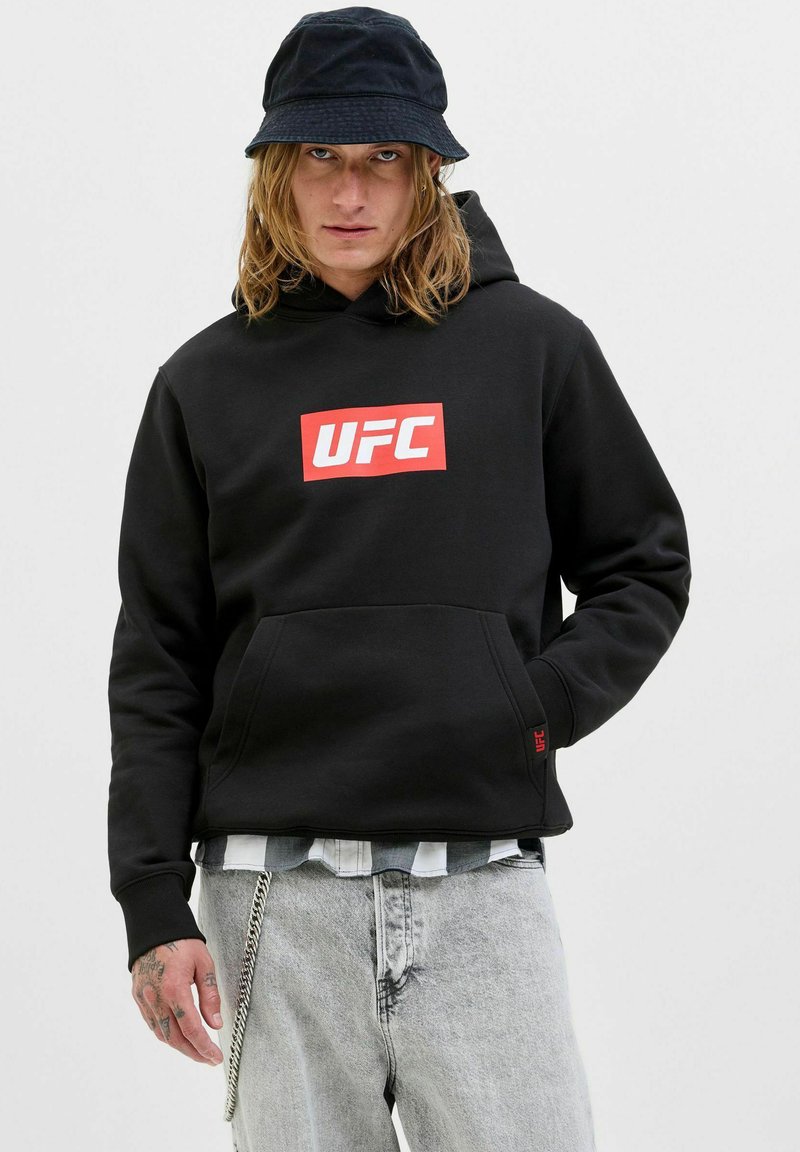Persona con felpa nera UFC, jeans chiari, cappello a secchiello nero, mano sinistra tatuata e accessorio a catena sui jeans.