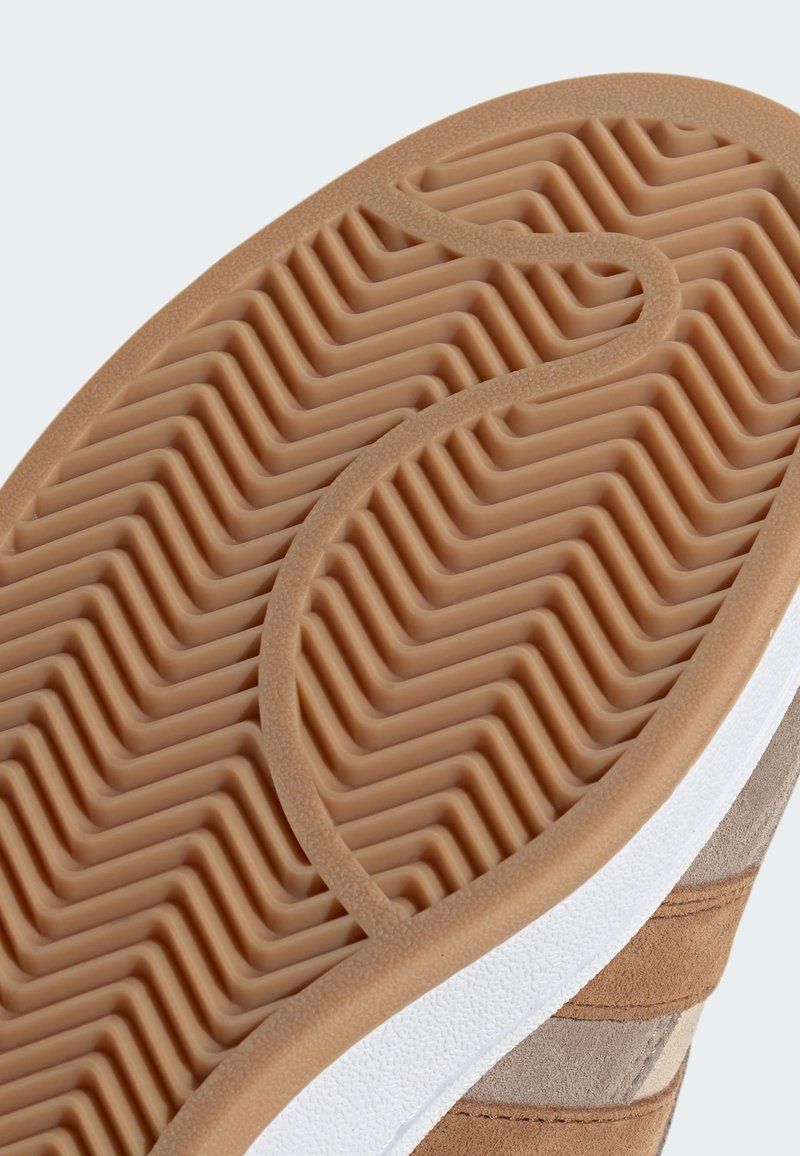 Suola di scarpa in gomma con un motivo a zigzag, caratterizzata da una superficie testurizzata color marrone chiaro, accentuata da un bordo bianco nella parte superiore.