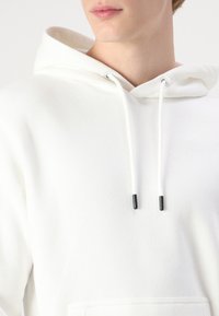 Hvid hættejakke med snøre i halsen, lavet af blødt materiale. Har sortspidset snøre og en forlomme. Minimalistisk design.