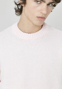 Pull en maille rose clair avec un col rond côtelé et une finition texturée. Le tissu semble doux et chaud, adapté à un port décontracté.