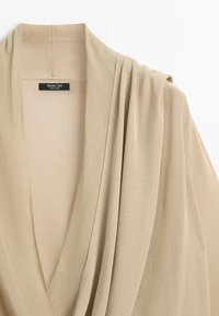 Béžový pletený cardigan s šálovým límcem a jemným efektem, označený Massimo Dutti, vyrobený v Číně, zobrazený na bílém pozadí.