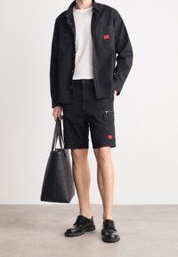 Man i svart skjortjacka, vit t-shirt, svarta cargoshorts med röda lappar, håller en svart tygväska och har på sig svarta läderskor med snörning.