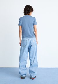 Osoba s kratkom kovrčavom kosom, odjevena u plavu majicu, labave svijetloplave traperice i crne tenisice, stoji na plavom podu okrenuta leđima.