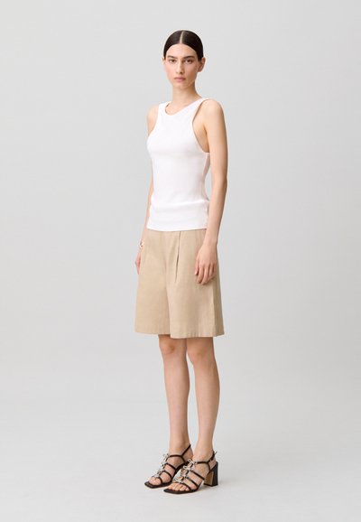 Mujer de pie, vestida con una camiseta sin mangas blanca, pantalones cortos beige hasta la rodilla y sandalias negras de tacón con detalles de lazo, frente a un fondo gris liso.