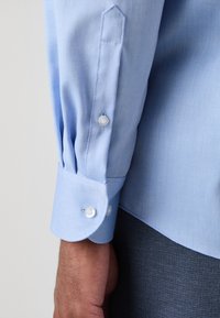 Lichtblauwe shirt met een textuurstof, knoopmanchetten en een puntige manchetdesign. De stof heeft een subtiel patroon en een gladde afwerking.