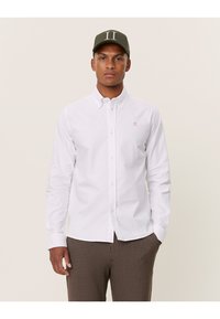 Camicia bianca con colletto a punta, dotata di un piccolo logo arancione sul petto, abbinata a pantaloni marroni a fantasia e un berretto verde.
