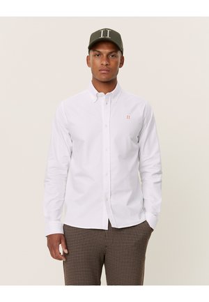 Camicia bianca con colletto a punta, dotata di un piccolo logo arancione sul petto, abbinata a pantaloni marroni a fantasia e un berretto verde.