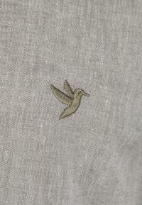 In Olivgrün auf leicht grauem, strukturiertem Stoff gestickter Kolibri, mit feinen Details in Flügeln und Körper.