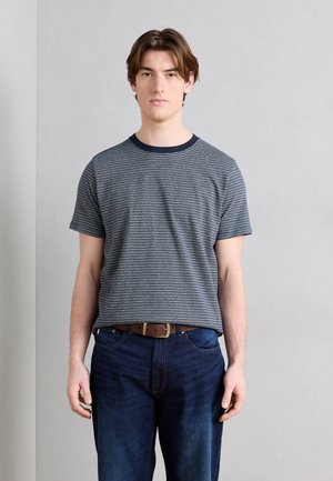 Junger Mann mit mittellangen braunen Haaren, der ein gestreiftes kurzärmeliges Hemd, einen braunen Gürtel und dunkelblaue Jeans vor einem schlichten Hintergrund trägt.