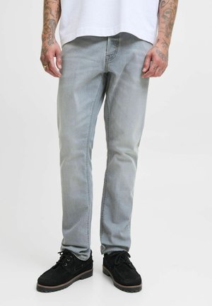 Man i ljusgrå jeans, svarta snörskor, vit t-shirt, med tatuerade armar och händer synliga mot en vit bakgrund.