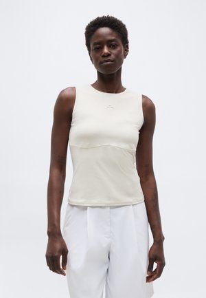 Ženska s kratkimi kodrastimi lasmi, oblečena v oprijeto belo majico brez rokavov in široke bele hlače, stoji pred enobarvno svetlo ozadje.