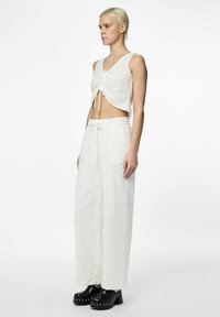 Witte crop top met details van een strik aan de voorkant, gecombineerd met losse, witte hoog-geplaatste broeken. Het model draagt zwarte chunky klompen. Gladde, lichte stof.