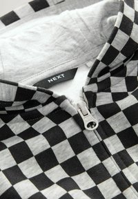 Svart och grå rutig zip-up hoodie med vit dragkedja och synlig "NEXT"-logotyp inuti kragen.