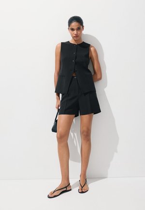 Femme portant un gilet noir sans manches boutonné, un short noir assorti, des sandales noires, et tenant un petit sac à main noir sur un fond blanc uni.