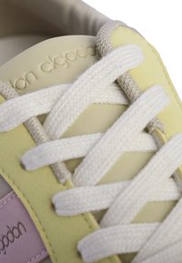Zapatilla de color amarillo claro con cordones blancos. Presenta un upper de cuero suave, un acento texturizado en rosa claro y un logotipo en relieve en la lengüeta.