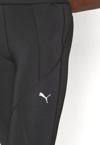 Svarta leggings med en slät textur, försedda med en vit Puma-logotyp, kontrasterande sömdetaljer och en åtsittande design för atletisk användning.