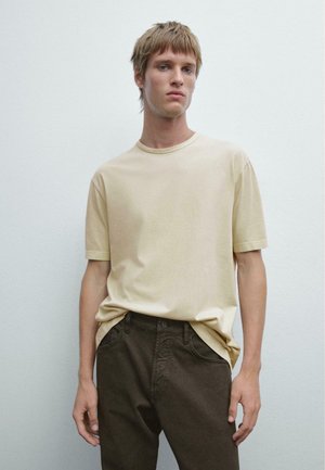 Beige bomulds t-shirt med rund halsudskæring og korte ærmer, parret med mørkebrune bukser med et simpelt og rent snit.
