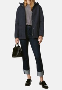 Giacca navy con cappuccio, maglione grigio a maglia e jeans scuri con risvolto chiaro. Una borsa nera e mocassini completano l'outfit.
