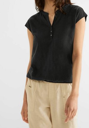 Kvinde iført en sort bluse med korte ærmer med tre knapper og beige høj-taljede bukser med forlommer, står mod hvid baggrund.