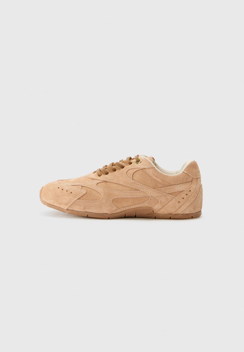 Tan suede sneaker with a curved design, rounded toe, and decorative stitching. Features a lace-up closure and textured rubber sole.