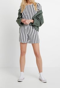 Conjunto a rayas en blanco y negro que incluye una camiseta de manga corta y pantalones cortos, complementado con una chaqueta verde oliva y zapatillas blancas.