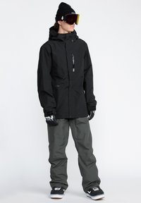 Veste de snowboard noire avec une fermeture éclair frontale, pantalon de neige gris, gants avec un design noir et blanc, et bottes de neige avec un accent blanc.