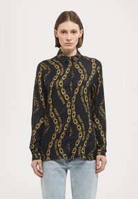 Camisa preta com um padrão de correntes douradas e texto do logotipo. Tem um decote de colarinho e mangas longas, confeccionada em tecido suave.