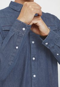 Mørkeblå denimskjorte med lange ærmer, med spredt krave, knapper foran og ved mansjettene, og et subtilt teksturert stoff.