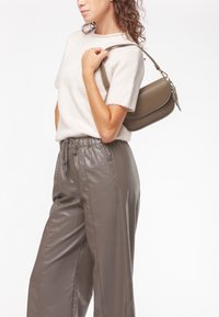 Borsa a tracolla in pelle verde oliva con forma curva, texture liscia e tracolla regolabile. Modella che indossa un maglione chiaro e pantaloni grigi.