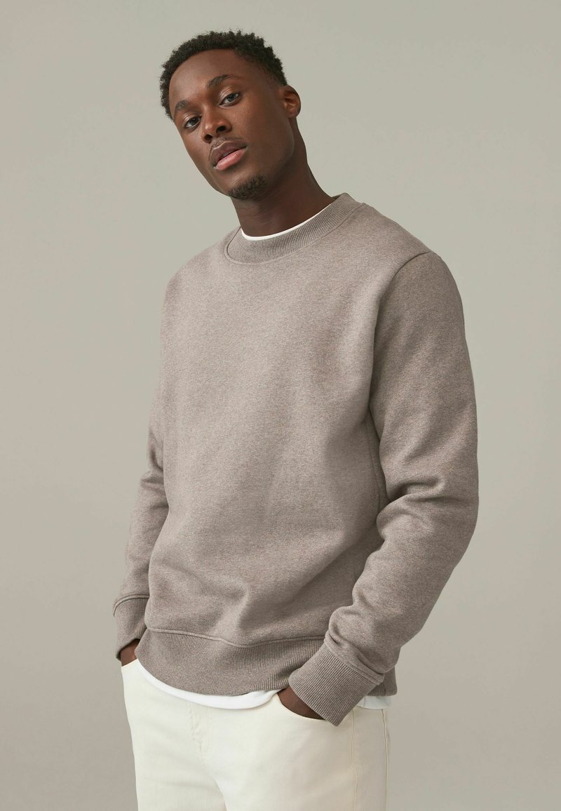 Sudadera gris de cuello redondo hecha de tela texturizada, con puños y dobladillo acanalados, llevada sobre una camisa blanca, combinada con pantalones de color claro.