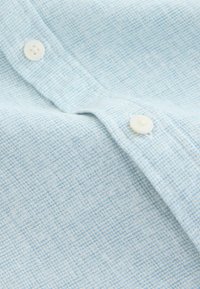 Chemise boutonnée bleu clair en tissu texturé, avec de petits boutons blancs et un motif à carreaux subtil.