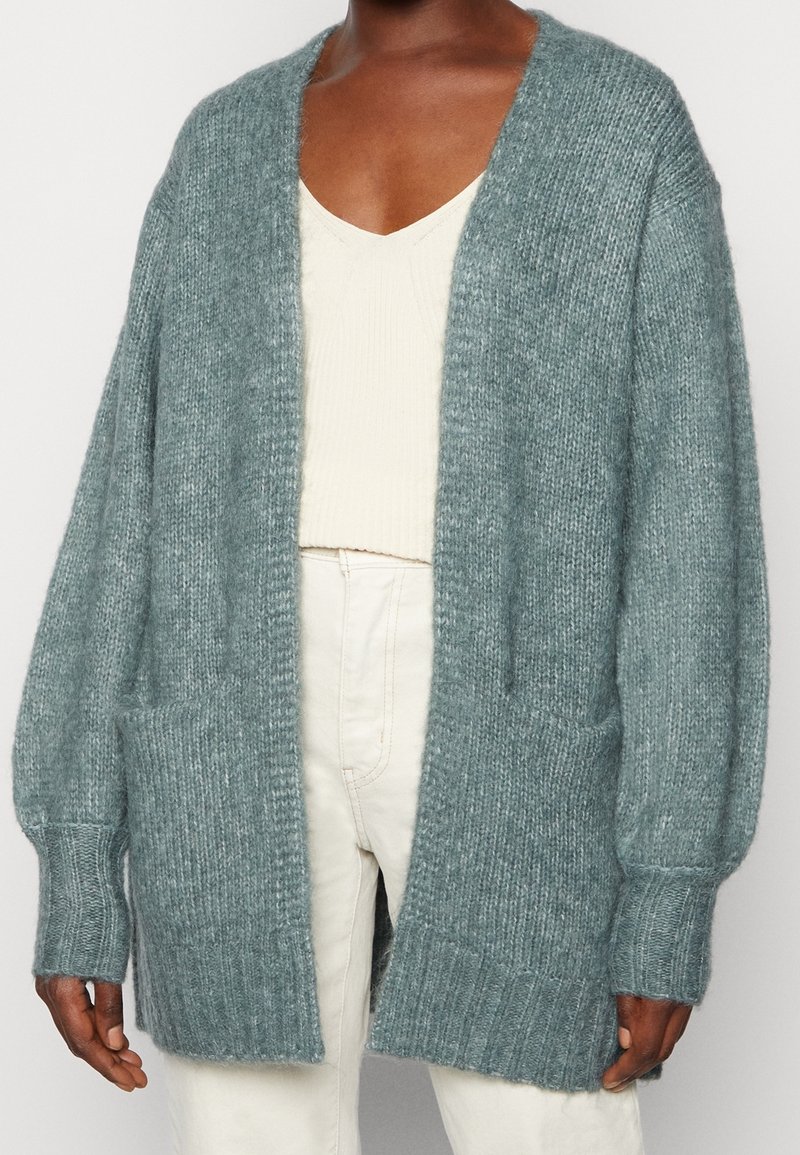 Lange, open voorkardigan in zachte teal gebreide stof. Beschikt over oversized mouwen, geribbelde manchetten, twee voorzakken en een relaxte pasvorm.