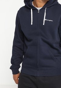 Felpa con cappuccio blu navy con zip, realizzata in misto cotone, con tasca a marsupio, coulisse bianche e logo Champion ricamato sul petto.