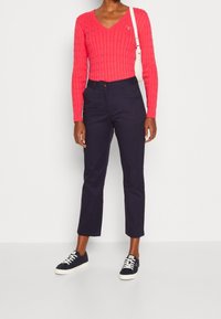 Žena v jasně růžovém svetr s pleteným vzorem a V-výstřihem, námořnických zkrácených kalhotách, černých teniskách s bílými tkaničkami a bílé kabelce.