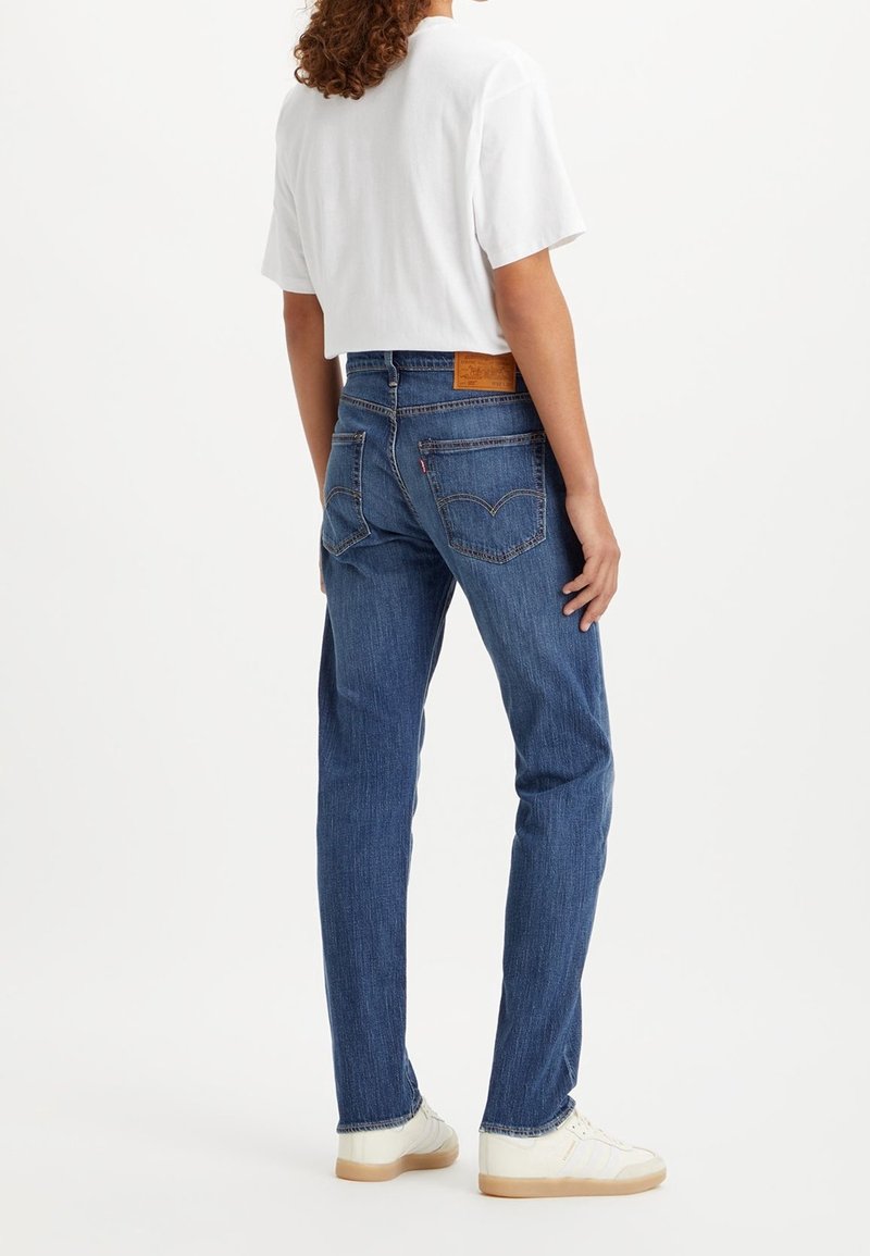 Personne portant un jean bleu à jambes droites et des baskets blanches, debout dos à la caméra, fond blanc uni.