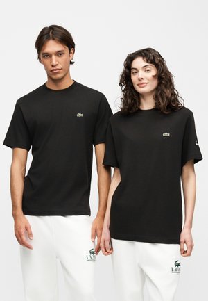 Deux mannequins portant des t-shirts noirs Lacoste et des pantalons blancs avec le logo vert Lacoste, debout côte à côte devant un fond uni.