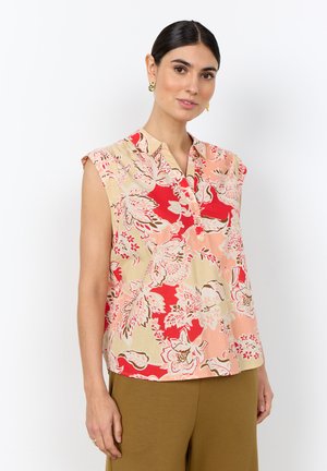 Femme aux cheveux foncés portant un chemisier sans manches à motifs floraux dans des tons rouges, beiges et roses, avec un pantalon vert olive et des boucles d'oreilles dorées.