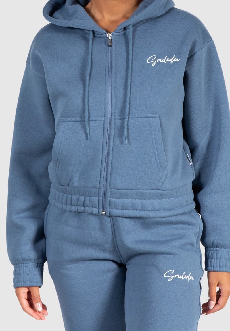 Femme portant un sweat à capuche bleu zippé et un pantalon de survêtement assorti, tous deux arborant des logos en texte cursif blanc sur la poitrine et la cuisse.