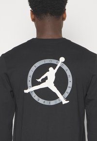 Musta pitkähiha t-paita, jossa on valkoinen grafiikka hyppivästä hahmosta ympyrämuotoisessa suunnittelussa, johon sisältyy "AIR JORDAN FLIGHT CLUB".